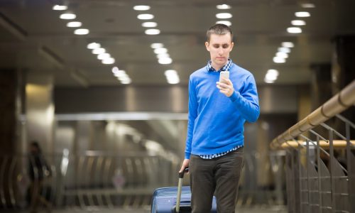 Portrait of young handsome guy wearing casual style clothes walking in modern station. Traveler making call using smartphone. Man travelling with suitcase making call, dialing number, sending text